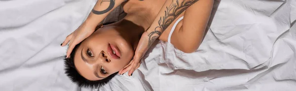Top view of young, tattooed and charming woman with short brunette hair touching face and looking away while laying on white bedding in studio, erotic photography, banner — Stock Photo