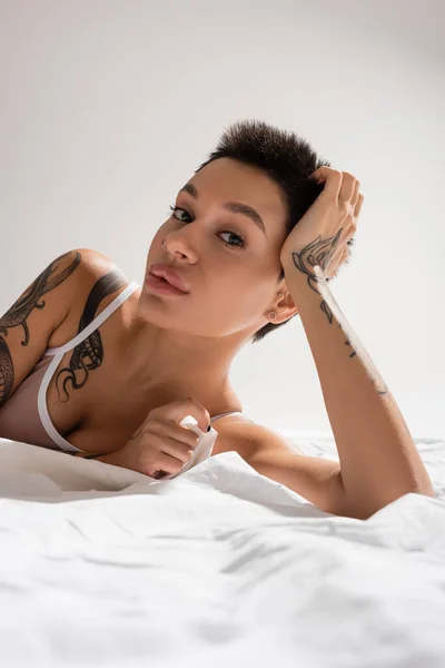 Jovem e atraente mulher em sutiã bege, com corpo tatuado sexy e cabelo curto morena posando em roupa de cama branca e olhando para a câmera em fundo cinza em estúdio, fotografia erótica — Fotografia de Stock