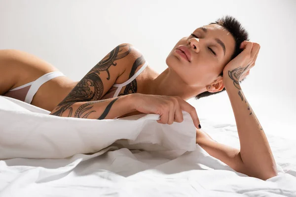 Mujer joven y sensual con pelo corto morena y cuerpo tatuado sexy posando en lencería con los ojos cerrados sobre ropa de cama blanca y fondo gris, fotografía erótica, arte de la seducción - foto de stock