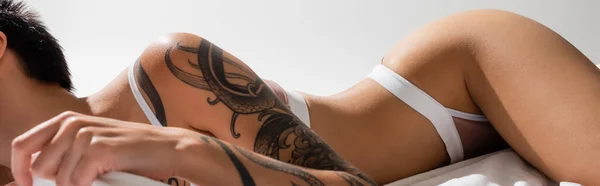 Vista parcial de mujer joven y sexy con cuerpo tatuado acostado en lencería en pose provocativa sobre ropa de cama blanca y fondo gris, fotografía erótica, pancarta - foto de stock