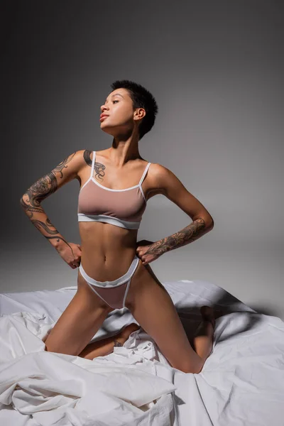 Full length of young, sexy and provocative woman with short brunette hair and tattooed body, wearing beige lingerie and posing on knees with hands on hips on white bedding and grey background — Stock Photo