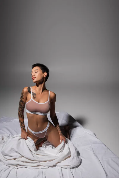 Mujer joven y provocadora con pelo corto morena y cuerpo sexy tatuado, vistiendo lencería beige y posando sobre rodillas sobre ropa de cama blanca y fondo gris, fotografía erótica - foto de stock