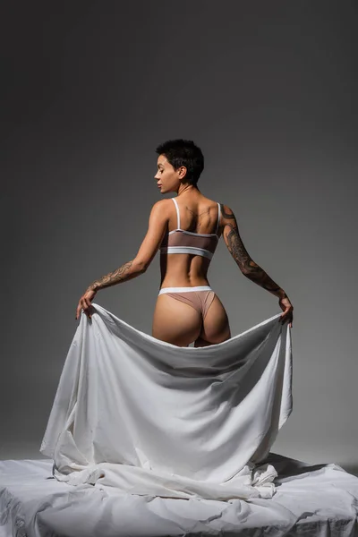 Back view de mulher deslumbrante e sexy em lingerie bege, com cabelo curto morena e corpo tatuado segurando lençol branco enquanto estava de pé sobre fundo cinza no estúdio, arte da sedução — Fotografia de Stock