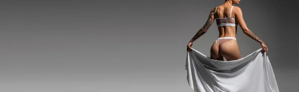 Visão parcial de mulher jovem e sexy com corpo tatuado e nádegas sexy em pé em lingerie bege enquanto posando com lençol branco no fundo cinza, fotografia erótica, banner — Stock Photo