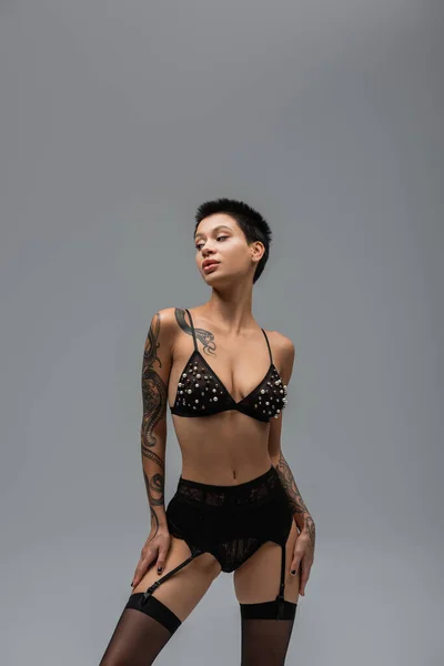 Mujer tatuada y expresiva con pelo corto morena y cuerpo tatuado posando en sujetador con perlas, bragas de encaje, liguero y medias negras y mirando hacia otro lado sobre fondo gris - foto de stock