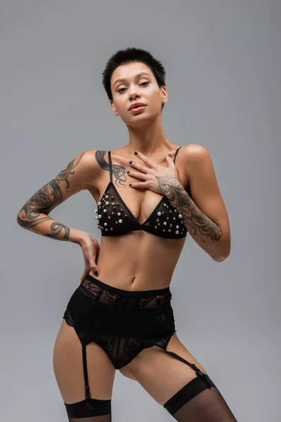 Mujer provocativa con cuerpo sexy tatuado y pelo corto morena, en sujetador con cuentas de perlas, bragas negras, liguero y medias posando con la mano en la cadera y mirando a la cámara sobre fondo gris - foto de stock