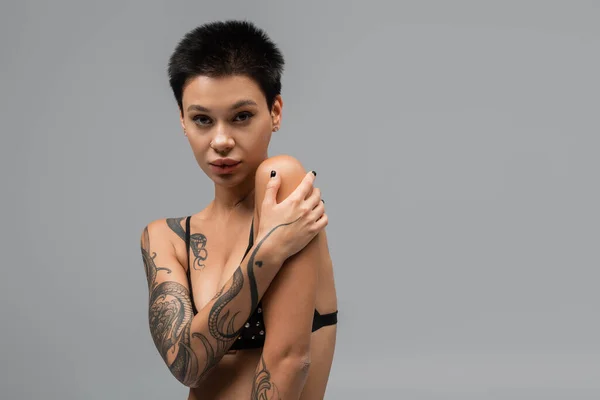 Young and passionate woman with short brunette hair and sexy tattooed body looking at camera while posing in black bra with pearl beads on grey background in studio, art of seduction — Stock Photo