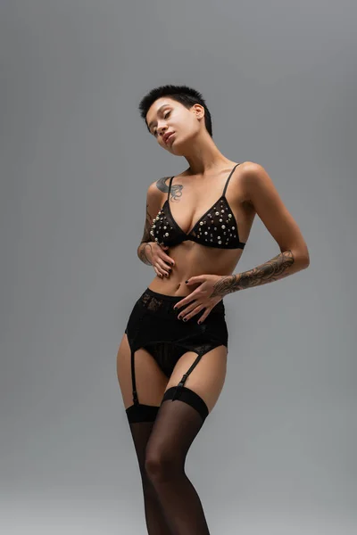 Passionate and stylish tattooed woman with short brunette hair posing in bra with pearl beads, black lace panties, garter belt and stocking on grey background in studio, erotic photography — Stock Photo