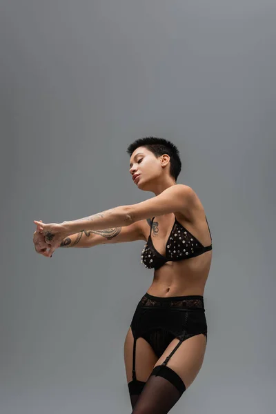 Young and graceful woman with sexy tattooed body and short brunette hair stretching arms and posing in black bra with pearl beads, lace panties, garter belt and stockings on grey background — Stock Photo
