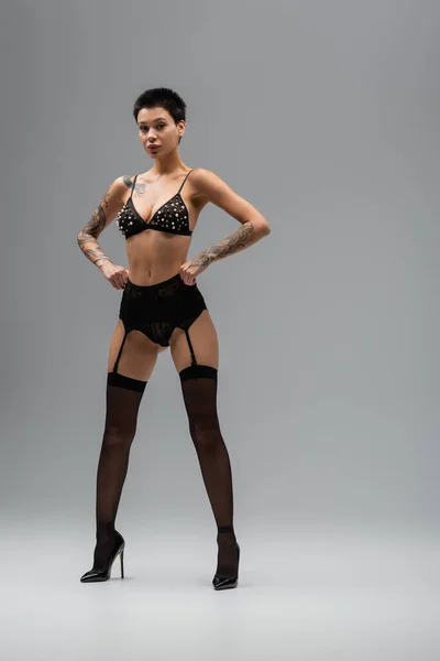 Full length of provocative woman with sexy tattooed body and short brunette hair standing with hands on hips in black bra with pearl beads, garter belt and black stockings on grey background — Stock Photo