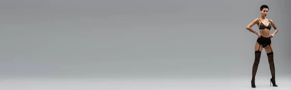 Volle Länge der sexy und selbstbewusste Frau mit schlankem tätowierten Körper, stehend in schwarzem BH mit Perlen, Spitzenhöschen, Strumpfgürtel, schwarzen Strümpfen und High Heels auf grauem Hintergrund, Banner — Stockfoto