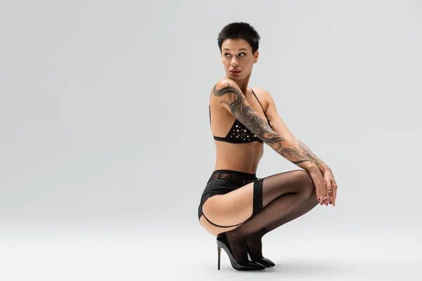 Full length of confident tattooed woman in bra with pearl beads, lace panties, garter belt, black stockings and high heels sitting on haunches and looking away on grey background, art of seduction — Stock Photo