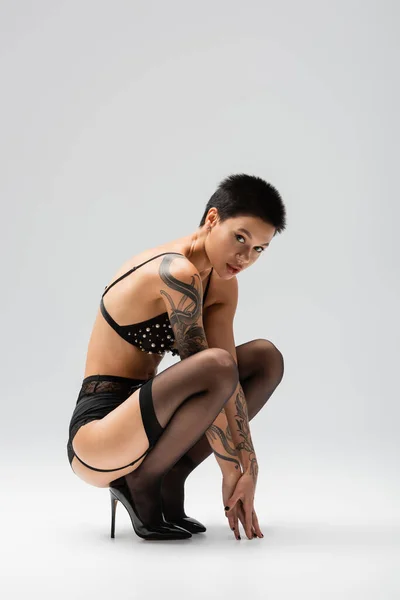 Full length of sexy woman with short brunette hair, in bra with pearl beads, garter belt, black stockings and high heels sitting on haunches and looking at camera on grey background, art of seduction — Stock Photo
