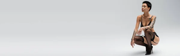 Volle Länge der jungen und begehrenswerten tätowierte Frau mit kurzen brünetten Haaren posiert in BH mit Perlen, Strumpfgürtel, schwarzen Strümpfen und High Heels auf grauem Hintergrund, Banner — Stockfoto