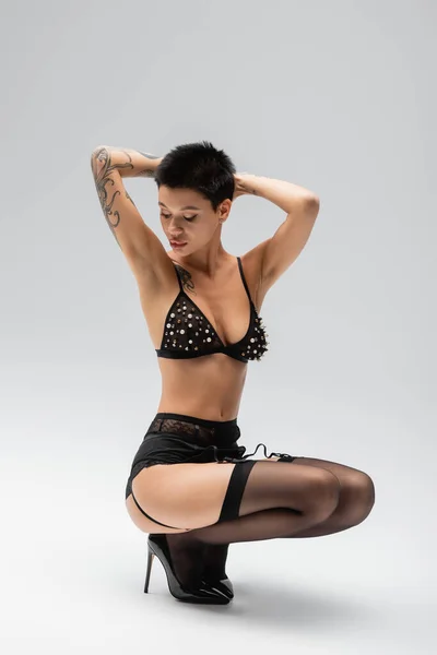 Full length of expressive tattooed woman in black stylish lingerie, bra with pearl beads, garter belt, black stockings and high heels posing on haunches with hands behind head on grey background — Stock Photo