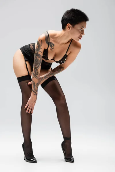 Comprimento total de mulher provocante e sexy com o corpo tatuado olhando para longe e posando em sutiã com pérolas, cinto de liga, meias pretas e saltos altos em fundo cinza, arte da sedução — Fotografia de Stock