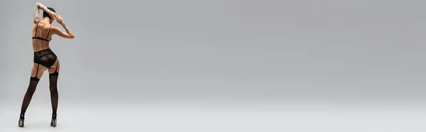 Vista posteriore della donna seducente tatuata con corpo sexy e capelli corti bruna in piedi in reggiseno, mutandine di pizzo, cintura giarrettiera, calze nere e tacchi alti su sfondo grigio, banner — Foto stock