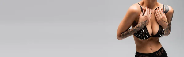 Vue partielle de femme sensuelle avec un corps tatoué sexy posant en culotte noire tout en touchant des bretelles de soutien-gorge avec des perles de perles sur fond gris, photographie érotique, bannière — Photo de stock