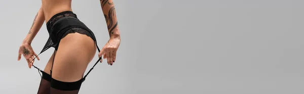 Vista recortada de mujer erótica joven con cuerpo tatuado sexy posando en bragas de encaje y medias negras mientras toca correas de liguero sobre fondo gris, arte de la seducción, pancarta - foto de stock