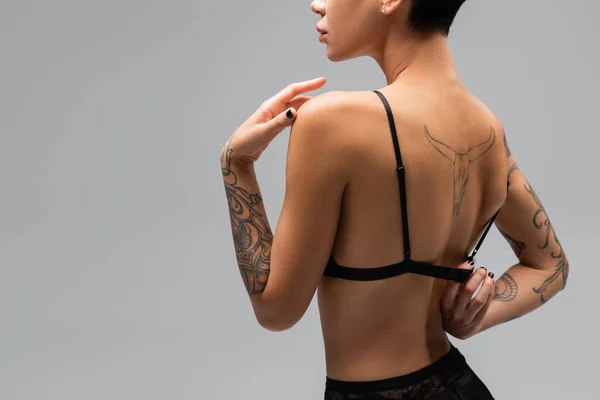 Visão traseira da jovem apaixonada com corpo tatuado sexy desabotoando sutiã preto enquanto posando em fundo cinza, arte da sedução, fotografia erótica, provocar — Fotografia de Stock