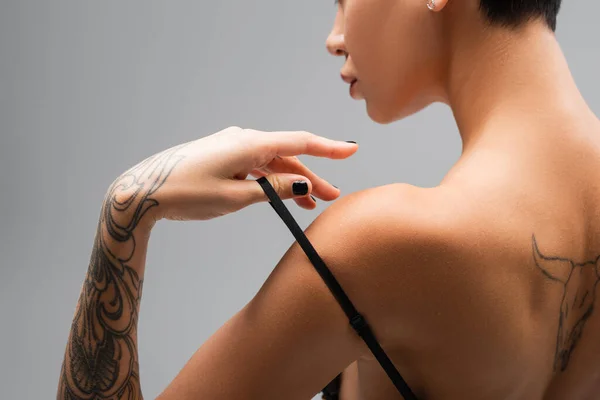 Vista parcial de mujer joven y apasionada con sexy cuerpo tatuado tocando correa de sujetador negro mientras posa sobre fondo gris, arte de la seducción, fotografía erótica - foto de stock