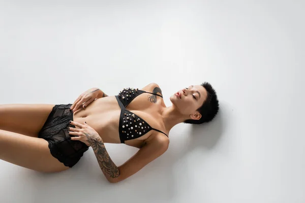 Visão de alto ângulo da mulher sensual e sexy com olhos fechados e corpo tatuado deitado em sutiã com pérolas e calcinha de renda preta em fundo cinza, fotografia erótica — Fotografia de Stock