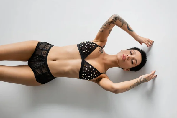 Вид зверху на молоду сексуальну жінку з татуйованим тілом і закритими очима в бюстгальтері з перламутровим бісером і мереживними трусиками, лежачи на сірому фоні, еротична фотографія — стокове фото