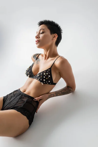 Молодая и сексуальная женщина с татуированным телом и короткими волосами брюнетки, сидящая в лифчике с жемчужными бусами и кружевными трусиками на сером фоне, эротическая фотография, искусство соблазнения — стоковое фото