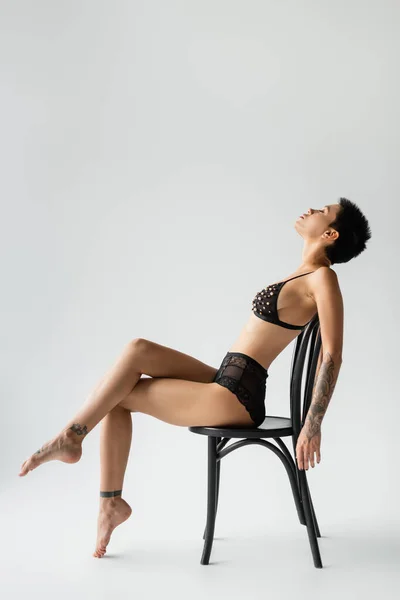 Vista lateral de mujer elegante con pelo corto morena y cuerpo tatuado sexy sentado en silla en sujetador negro con cuentas de perlas y bragas de encaje sobre fondo gris, fotografía erótica - foto de stock