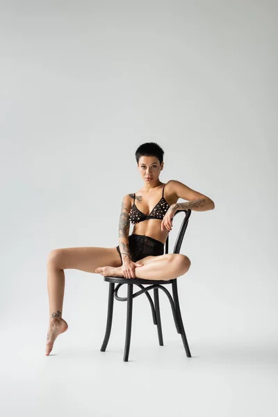 Full length of young, sexy and provocative woman with tattooed body and short brunette hair sitting on black chair in bra with pearl beads and lace panties on grey background — Stock Photo