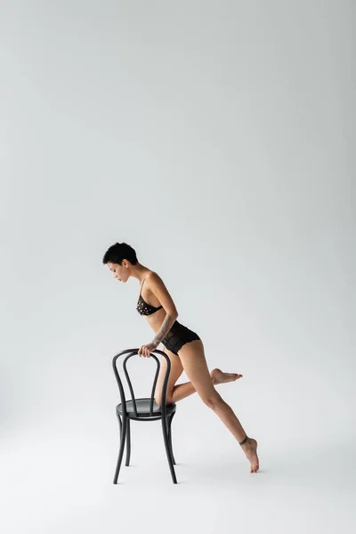 Volle Länge der glamourösen und tätowierten Frau mit sexy Körper und kurzen brünetten Haaren posiert mit schwarzem Stuhl in BH mit Perlen und Spitzenhöschen auf grauem Hintergrund — Stockfoto