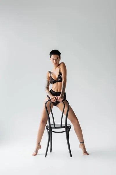 Full length of young seductive woman with short brunette hair and sexy tattooed body, in bra with pearl beads and lace panties looking at camera while posing with chair on grey background — Stock Photo