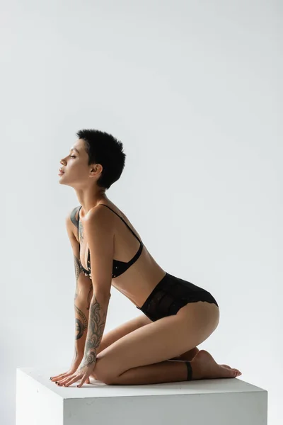 Full length of charming and sexy woman with short brunette hair and tattooed body posing on white cube in black bra with pearl beads and lace panties on grey background — Stock Photo