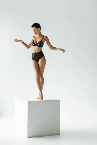 Full length of appealing woman with sexy tattooed body and short brunette hair standing on white cube in black bra with pearl beads and lace panties on grey background — Stock Photo