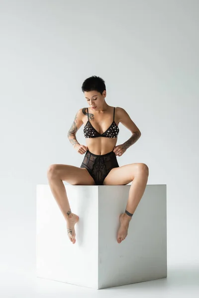 Full length of provocative and sexy woman with tattooed body and short brunette hair wearing bra with pearl beads and adjusting black lace panties while sitting on white cube on grey background — Stock Photo