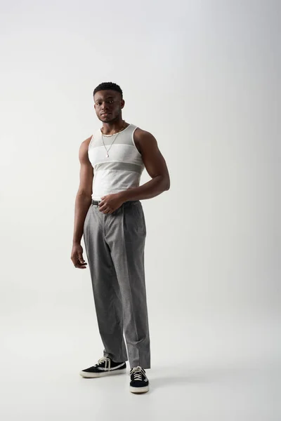 Full length of young afroamerican man in sleeveless t-shirt and pants looking at camera while standing on grey background, contemporary shoot featuring casual attire — Stock Photo