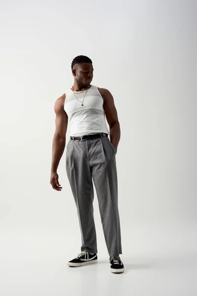 Comprimento total de jovem afro-americano em camiseta sem mangas e calças de mão no bolso e em pé sobre fundo cinza, tiro contemporâneo com trajes elegantes — Fotografia de Stock