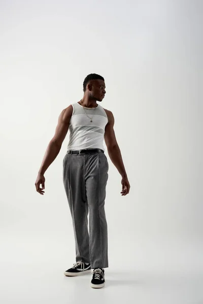 Piena lunghezza di giovane afroamericano alla moda in t-shirt senza maniche e pantaloni guardando altrove su sfondo grigio, servizio contemporaneo con abbigliamento casual — Foto stock