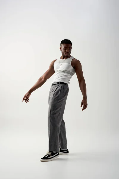 Piena lunghezza di moda giovane uomo afroamericano in pantaloni, t-shirt senza maniche e scarpe da ginnastica in piedi su sfondo grigio, servizio fotografico contemporaneo con abbigliamento casual — Foto stock