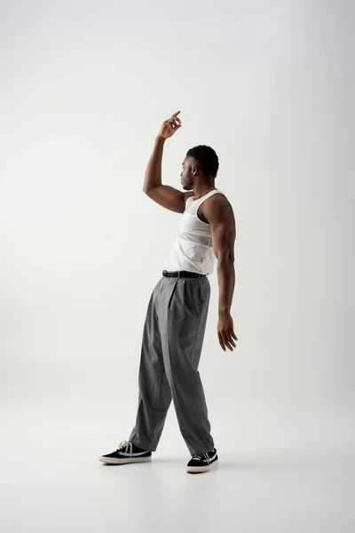 Вид збоку на м'язистого молодого афроамериканця в без рукавів футболці та штанях, що дивиться на сірий фон, сучасна стрілянина з повсякденним вбранням — стокове фото