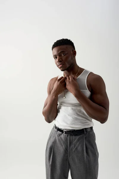 Homem americano africano musculoso e jovem em t-shirt sem mangas e calças tocando colar e olhando para a câmera isolada em cinza, tiro contemporâneo com traje casual — Fotografia de Stock