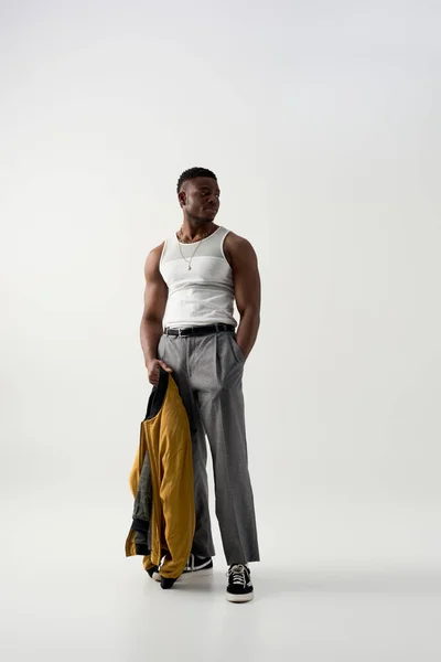 Full length of trendy young african american man in sleeveless t-shirt and pants holding bomber jacket on grey background, contemporary shoot featuring casual attire — Stock Photo