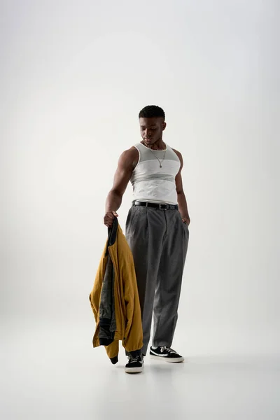 Full length of trendy african american man in tank top and pants looking at bomber jacket on grey background, contemporary shoot featuring stylish attire, muscular — Stock Photo