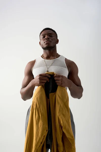 Visão de baixo ângulo de confiante jovem afro-americano em top tanque segurando jaqueta de bombardeiro isolado em cinza, tiro contemporâneo com traje elegante, muscular — Fotografia de Stock