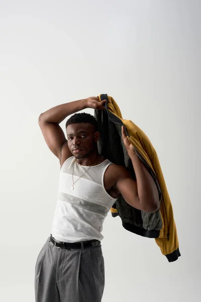 Уверенный молодой афроамериканец в майке, одет в бронежилет и стоит изолированный на сером, современном снимке со стильной одеждой, мускулистый, модный вперед — стоковое фото