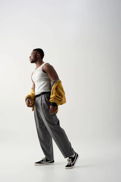 Side view of muscular african american man in bomber jacket and sleeveless t-shirt standing on grey background, contemporary shoot featuring stylish attire, fashion statement — Stock Photo