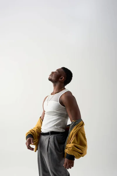 Вид сбоку на сильного и модного молодого африканского мужчину в майке танка и пиджаке бомбардировщика, стоящего изолированно на серой, современной съемке со стильной одеждой, мускулистый — стоковое фото