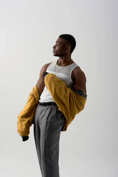 Seitenansicht des gut aussehenden afroamerikanischen Models in Bomberjacke und ärmellosem T-Shirt posiert isoliert auf grauem, zeitgenössischem Shooting mit stylischer Kleidung, muskulös — Stockfoto