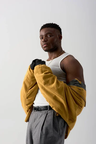 Портрет молодого и уверенного африканского мужчины в куртке подрывника и футболке без рукавов, стоящей изолированной на серой, современной съемке со стильной одеждой, мускулистой — стоковое фото
