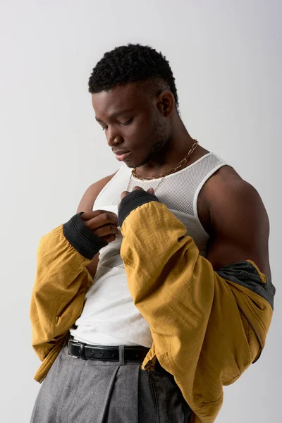 Young and handsome african american man in bomber jacket and sleeveless t-shirt touching necklace isolated on grey, contemporary shoot featuring stylish attire, muscular — Stock Photo
